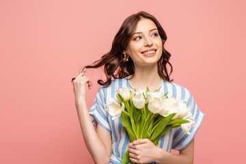 happy girl touching hair while holding bouquet of white tulips and looking away isolated on pink