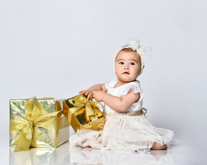 Fototapeta premium Baby girl in dress and headband, barefoot. She holding golden gift box, sitting on floor isolated on white. Close up, copy space