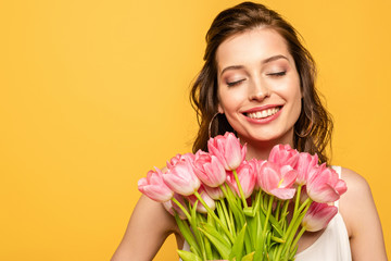 young woman smiling with closed eyes while holding bouquet of pink tulips isolated on yellow