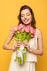 happy young woman holding bouquet of pink tulips while smiling with closed eyes isolated on yellow