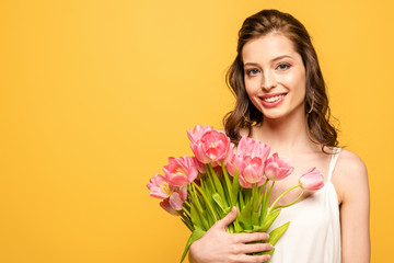 cheerful young woman smiling at camera while holding bouquet of pink tulips isolated on yellow