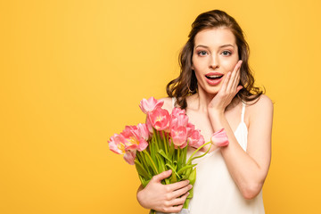shocked young woman holding bouquet of pink tulips and touching face while looking at camera isolated on yellow