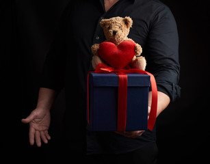 adult man in a black shirt holds a blue square box tied with a red ribbon and sits on top of a brown teddy bear