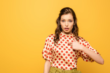 shocked young woman pointing with finger at herself while looking at camera isolated on yellow