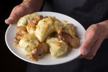Man holds a plate with varenyky or dumplings, with meat, onions and bacon on a white plate. Pierogi, dumplings served with caramelized salted onion in bowl. Male in a black apron without face. close-u