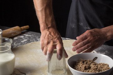 A man is making a dough for varenyky or dumplings, with meat, onions and bacon. Man in a black apron without face. Man squeezes out circles of dough with a glass. Making dough by male hands at bakery 