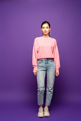 full length view of pretty, confident girl looking at camera while standing on purple background