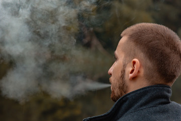 A guy with a beard sits on the lake and smokes an electronic cigarette. Blows puffs of smoke. The concept of unity with nature. Rest near the water. Vape, Man Vaping. lips and smoke, a lot of smoke.