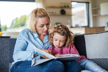 A cute small girl with mother on sofa indoors at home, reading book.