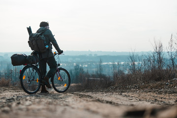 Fototapeta premium A man with a backpack and a bicycle stands and looks into the distance. hilltop overlooking a valley in haze, a city on the horizon. winter or autumn landscape road. Things, a tent on the trunk.