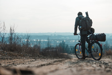 A man with a backpack and a bicycle stands and looks into the distance. hilltop overlooking a valley in haze, a city on the horizon. winter or autumn landscape road. Things, a tent on the trunk.