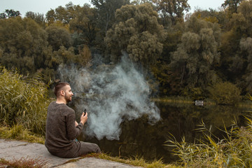 A guy with a beard sits on the lake and smokes an electronic cigarette. Blows puffs of smoke. The concept of unity with nature. Rest near the water. Vape, Man Vaping. lips and smoke, a lot of smoke.