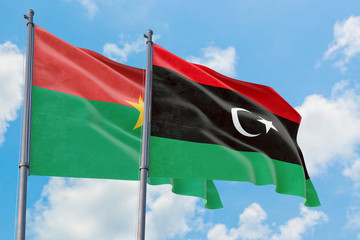 Libya and Burkina Faso flags waving in the wind against white cloudy blue sky together. Diplomacy...
