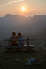 Boy and a girl sit hugging each other on a bench and watch the sunset nere winding road seen from Sedlo Pass in Durmitor National Park in Montenegro at sunset