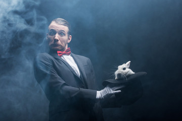 emotional shocked magician holding white rabbit in hat, dark room with smoke