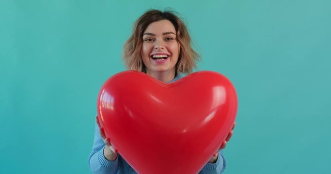 Valentine's Day cheerful girl catches red heart inflatable ball, presses it to him, smiles happily looking at camera blue background. Women, symbol of love. Holiday of all lovers. Feelings, emotions