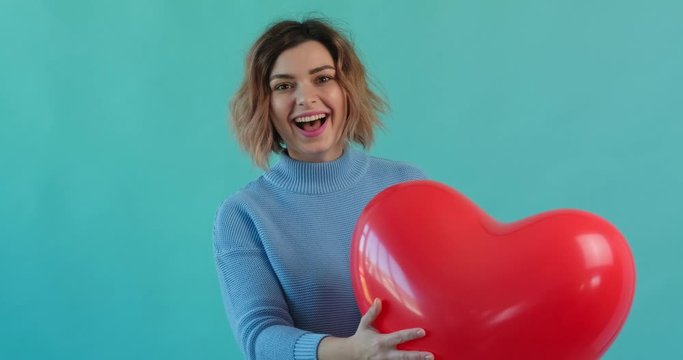 Valentine's day cheerful girl throws up red heart balloon, presses it to herself, smiling happily looking at camera blue background. Woman, symbol of love. Holiday of all lovers. Feelings, emotions