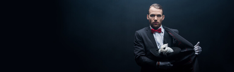 panoramic shot of professional magician holding white rabbit in hat, dark room with smoke