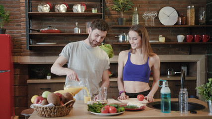 Handsome smiling bearded Caucasian man pours fresh orange juice for his sexy hot woman in glasses on modern kitchen background. Couple goals, taking care, healthy food, organic nutrition. Close up