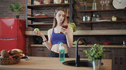 Gorgeous fit young Caucasian sportswoman in stylish wear standing at the kitchen table, eating green apple and using smartphone. Fitness, sports app, healthy lifestyle, nutrition. Close up view