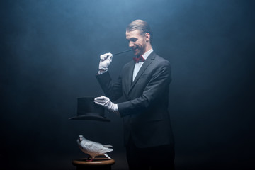 joyful magician showing trick with dove, wand and hat in dark room with smoke