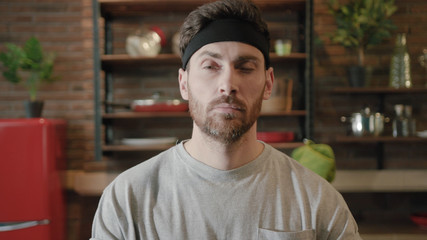 Millennial male practicing yoga meditation with eyes closed at home, then shows up his tongue. Bearded man in casual taking rest for wellness, stress control and mental health. Positive emotions