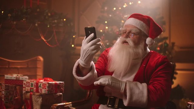 Santa Claus uses a smartphone and talks via video link while conducting a conference. Santa Claus and selfie camera, online broadcasts.