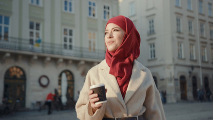 Obraz premium Attractive middle eastern woman in red hijab holding coffee cup walking in the beautiful street in the city. Portrait of cheerful Muslim girl admiring her vacation.