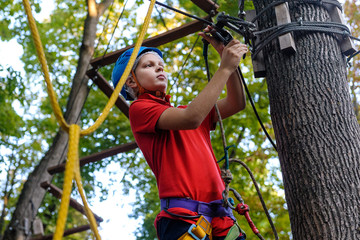 A teenage boy climbs in a rope park. There is a protective helmet on his head. The boy has special climbing equipment. Summer.