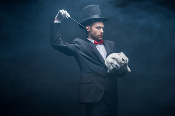 emotional magician in suit and hat showing trick with wand and white rabbit, dark room with smoke