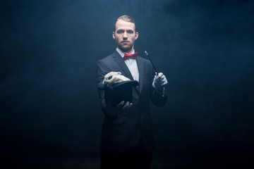 serious professional magician in suit showing trick with wand and white rabbit in hat, dark room with smoke