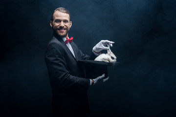 cheerful magician in suit showing trick with white rabbit in hat, dark room with smoke - obrazy, fototapety, plakaty