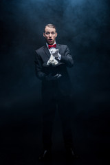 excited magician in suit showing trick with white rabbit in hat, dark room with smoke