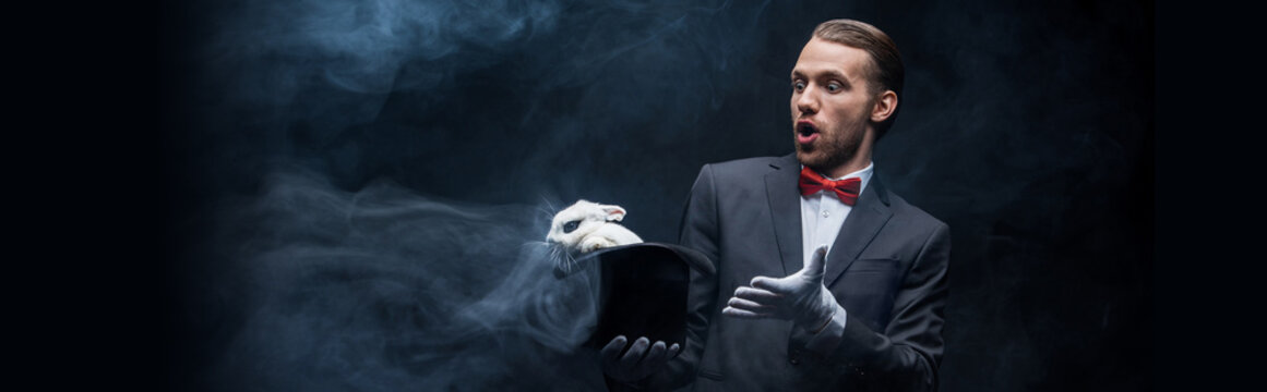 panoramic shot of shocked magician in suit holding hat with white rabbit, dark room with smoke