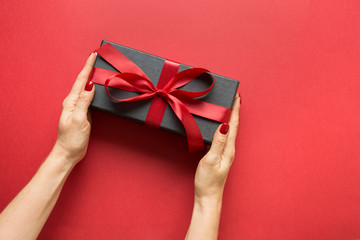 Woman's hands holding black gift box wrapped with red ribbon on red surface. Valentine day card.