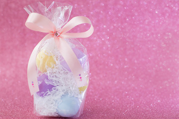 Multi-colored purple, pink, blue, yellow Easter eggs in a transparent gift package, decorated with a bow and rhinestones, on a pink shiny bokeh background. Symbol of celebration of religious holiday