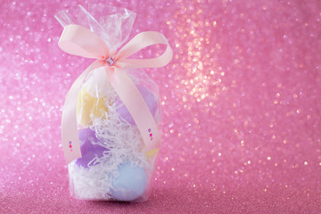 Multi-colored purple, pink, blue, yellow Easter eggs in a transparent gift package, decorated with a bow and rhinestones, on a pink shiny bokeh background. Symbol of celebration of religious holiday