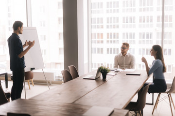 Young businessman presenting new project to colleagues on whiteboard. Confident employee giving presentation to partners in modern office room against large window. Concept of office life.