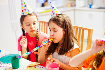 Little pretty girl trying to blow a whistle at party. Sisters sitting at birthday table