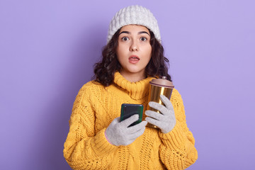 Close up indoor photo of surprised magnetic young brunette opening mouth widely, looking directly at camera, holding smartphone and thermal mug in both hands, having shocked facial expression.