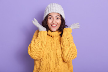 Horizontal picture of beautiful tender emotional young lady wearing grey hat, gloves and yellow sweater, raising arms, looking directly at camera, laughing sincerely. People and emotions concept.