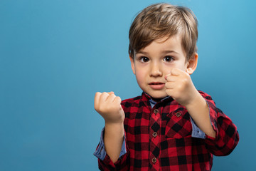 Portrait of small little caucasian boy young child kid standing in front of the blue wall background wearing red and black shirt looking to camera in studio holding fists in fighting stance position