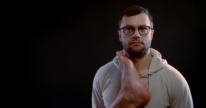 a young heavily unshaven brunette with glasses stands on a black background. a big man's bare foot rises from the bottom and slaps the guy on his cheek