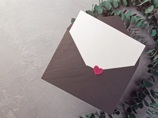 A blank sheet of paper is in a black envelope on a gray background. Decoration with eucalyptus and heart figure. Flat lay, top view. Copy space.