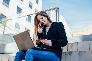 Attractive young woman with long brown hair shopping online with her smartphone and laptop while holding a credit card