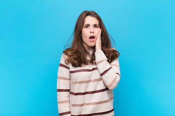 young pretty woman feeling shocked and astonished holding face to hand in disbelief with mouth wide...
