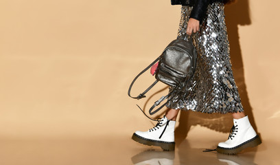 Cropped photo of a girl in a shiny skirt, white shoes. Posing goes sideways with a silver backpack on a beige background. Hipster style, fashion, beauty.