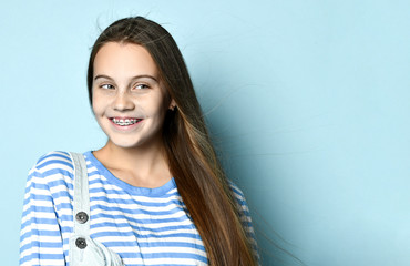 Teenage girl in jeans overall and striped sweatshirt. She is smiling while posing against blue...