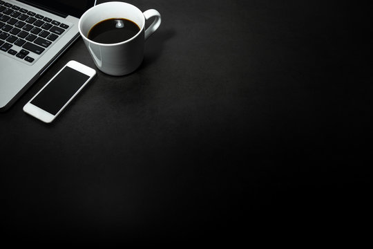 Laptop, white mobile phone  and a cup of black coffee against empty dark background, working office theme