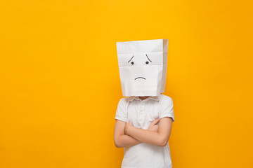 Cute little school boy standing with a paper bag on his head - sad face on yellow background,...